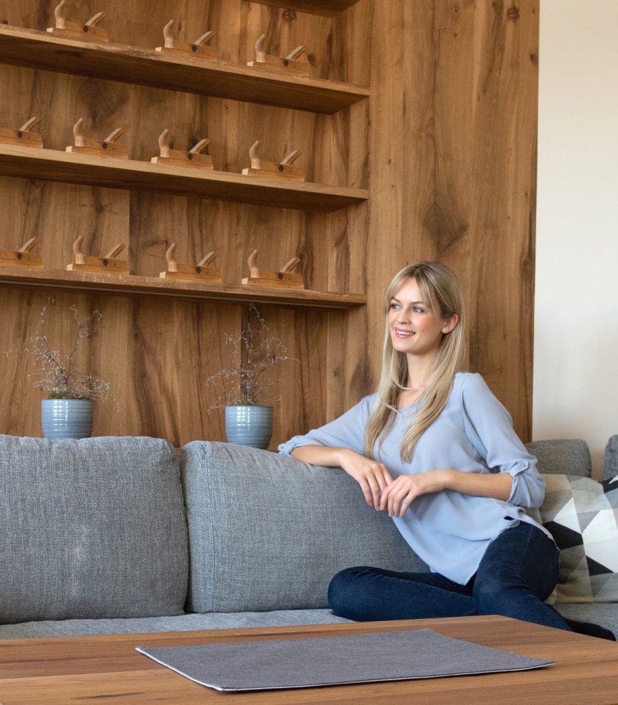 Eine Frau sitzt auf dem Sofa, im Hintergrund eine vom Tischler gefertigte Wand aus Holze mit Hobel.