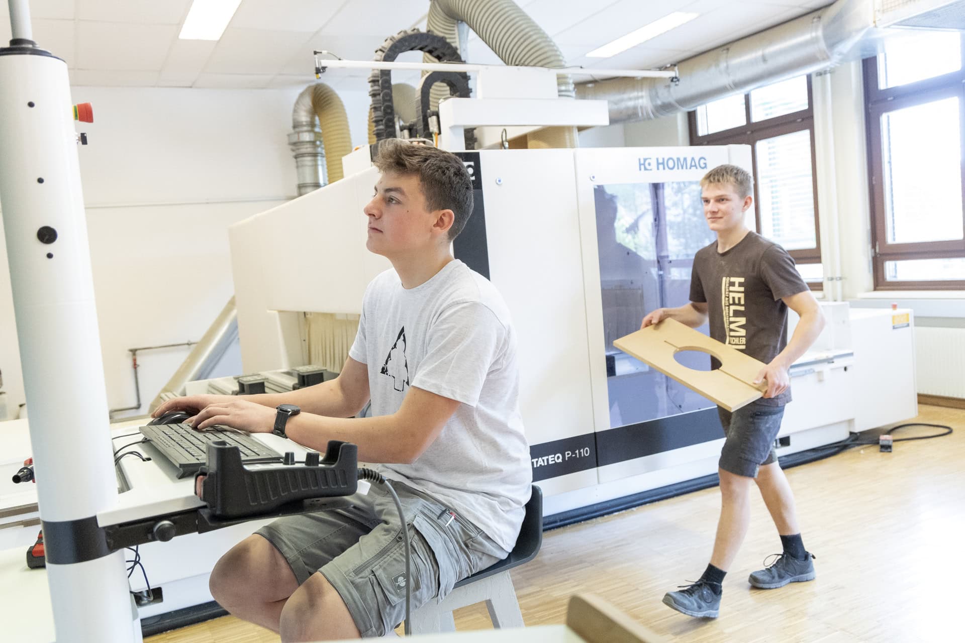 LLWB-tischler25-maschinen-001 Zwei junge Männer arbeiten in einer hellen Tischlerwerkstatt an einer CNC-Maschine. Im Vordergrund sitzt ein Auszubildender am Computer und steuert die Anlage, während im Hintergrund ein Kollege eine zugeschnittene Holzplatte mit runder Aussparung trägt. Große Absaugrohre und die Maschine sind sichtbar, Tageslicht fällt durch die Fenster.