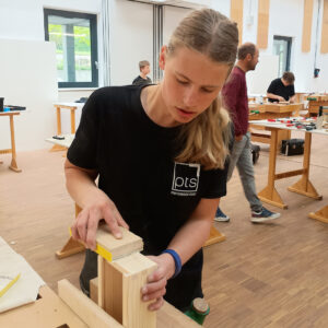 Junge Teilnehmerin schleift konzentriert ein Holzwerkstück in einer Tischlerei-Werkstatt während eines Wettbewerbs.