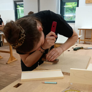 Junge Teilnehmerin arbeitet konzentriert an einem Holzstück in einer Tischlerei-Werkstatt während eines Wettbewerbs.