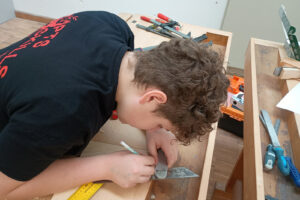 Junger Teilnehmer markiert präzise ein Holzstück mit Winkel und Bleistift an einer Werkbank während eines Tischlerei-Wettbewerbs.