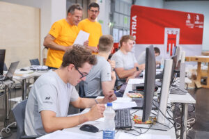 Teilnehmer eines Tischler-Wettbewerbs arbeiten an Computern und Unterlagen, während Jury im Hintergrund die Aufgaben überwachen.