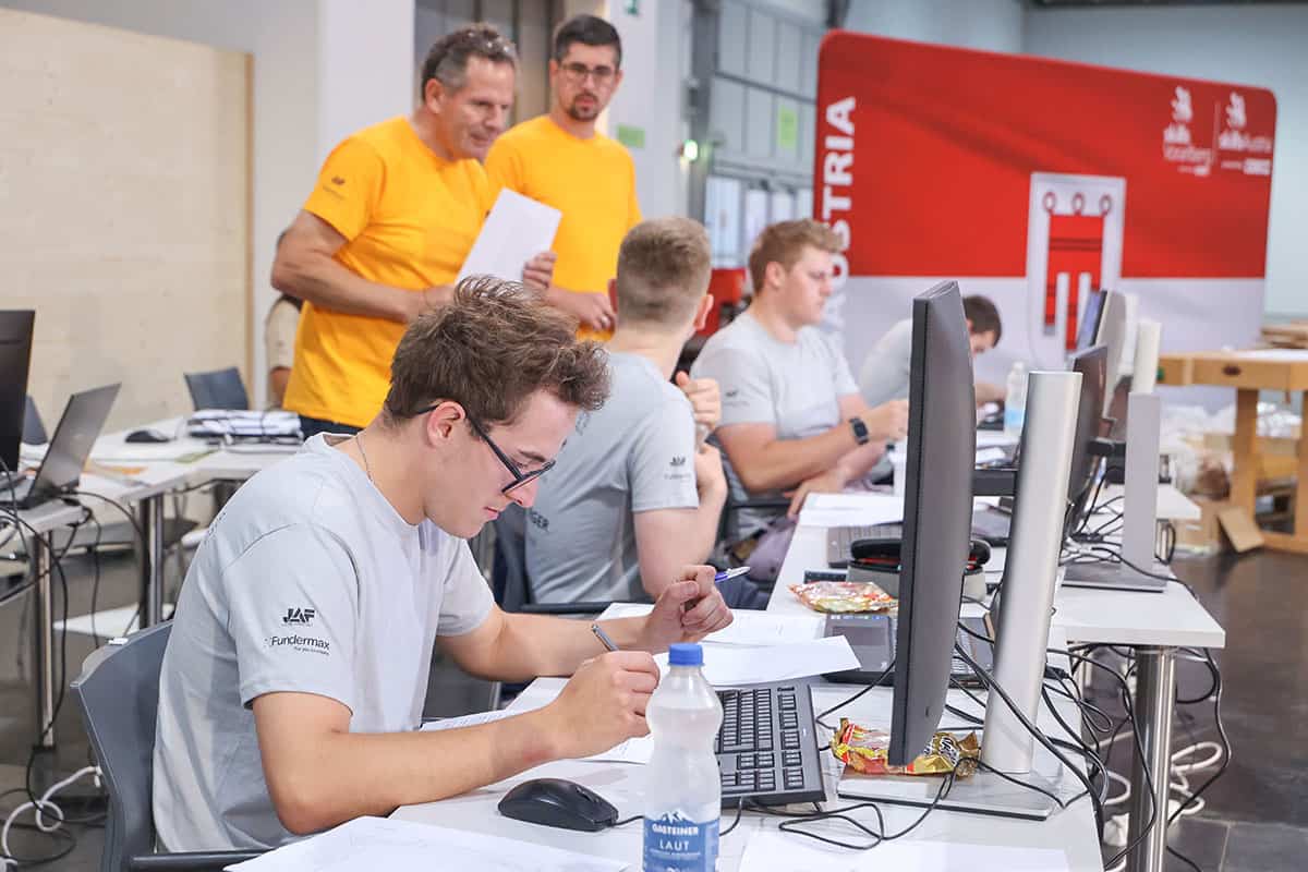 tischler-bundeslehrlingswettbewerb-2026-04 Teilnehmer eines Tischler-Wettbewerbs arbeiten an Computern und Unterlagen, während Jury im Hintergrund die Aufgaben überwachen.