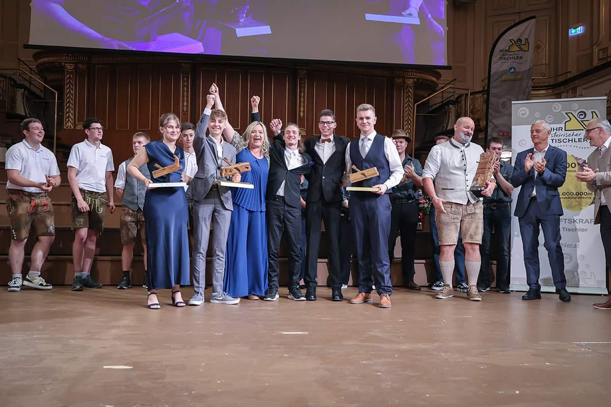 Siegerehrung eines Tischler-Lehrlingswettbewerbs auf einer Bühne. Mehrere junge Teilnehmerinnen und Teilnehmer halten Trophäen aus Holz, während die Gewinner in der Mitte jubelnd die Arme heben. Um sie herum stehen Organisatoren und weitere Teilnehmende, einige applaudieren. Im Hintergrund sind Banner der Tischler-Innung und eine große Leinwand zu sehen.