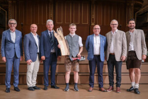 Junger Tischler-Lehrling steht mit einer großen Holz-Trophäe und einem Werkzeugpreis auf einer Bühne, umgeben von mehreren Gratulanten in Anzügen und Tracht.