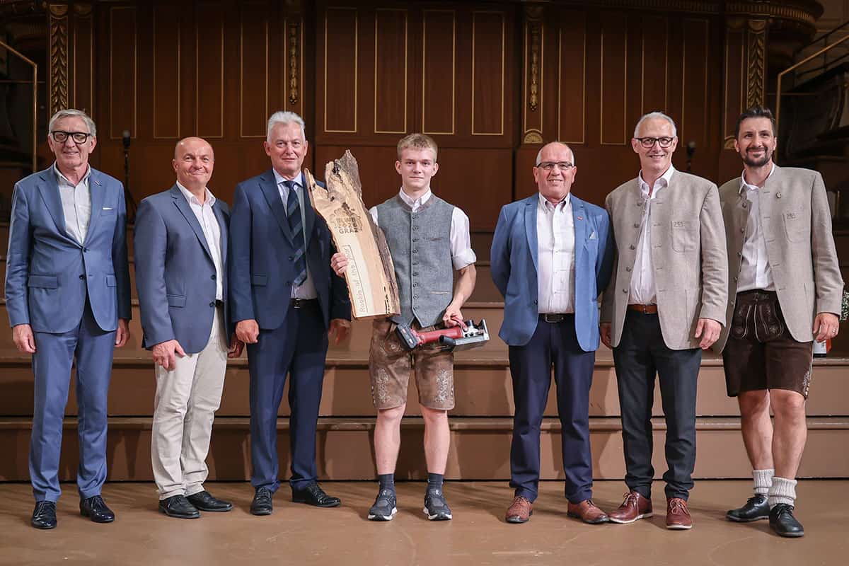 tischler-bundeslehrlingswettbewerb-2026-jakob-riedler-rookie-of-the-year-2026 Junger Tischler-Lehrling steht mit einer großen Holz-Trophäe und einem Werkzeugpreis auf einer Bühne, umgeben von mehreren Gratulanten in Anzügen und Tracht.