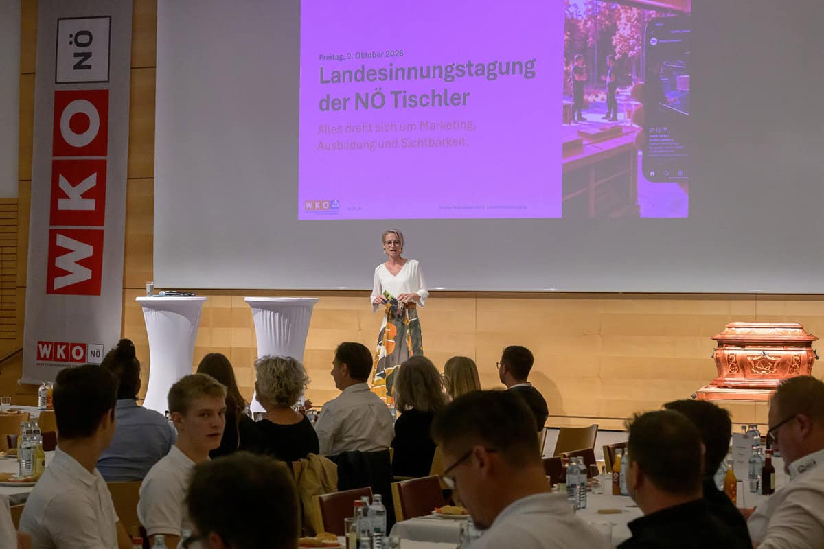 Landesinnungstagung 2026 – „Miteinander“ im Mittelpunkt Eine Referentin steht auf der Bühne vor einer Präsentation zur „Landesinnungstagung der NÖ Tischler“ am 2. Oktober 2026. Im Vordergrund sitzen zahlreiche Teilnehmerinnen und Teilnehmer an gedeckten Tischen. Links ist ein großes WKO-Niederösterreich-Banner zu sehen.